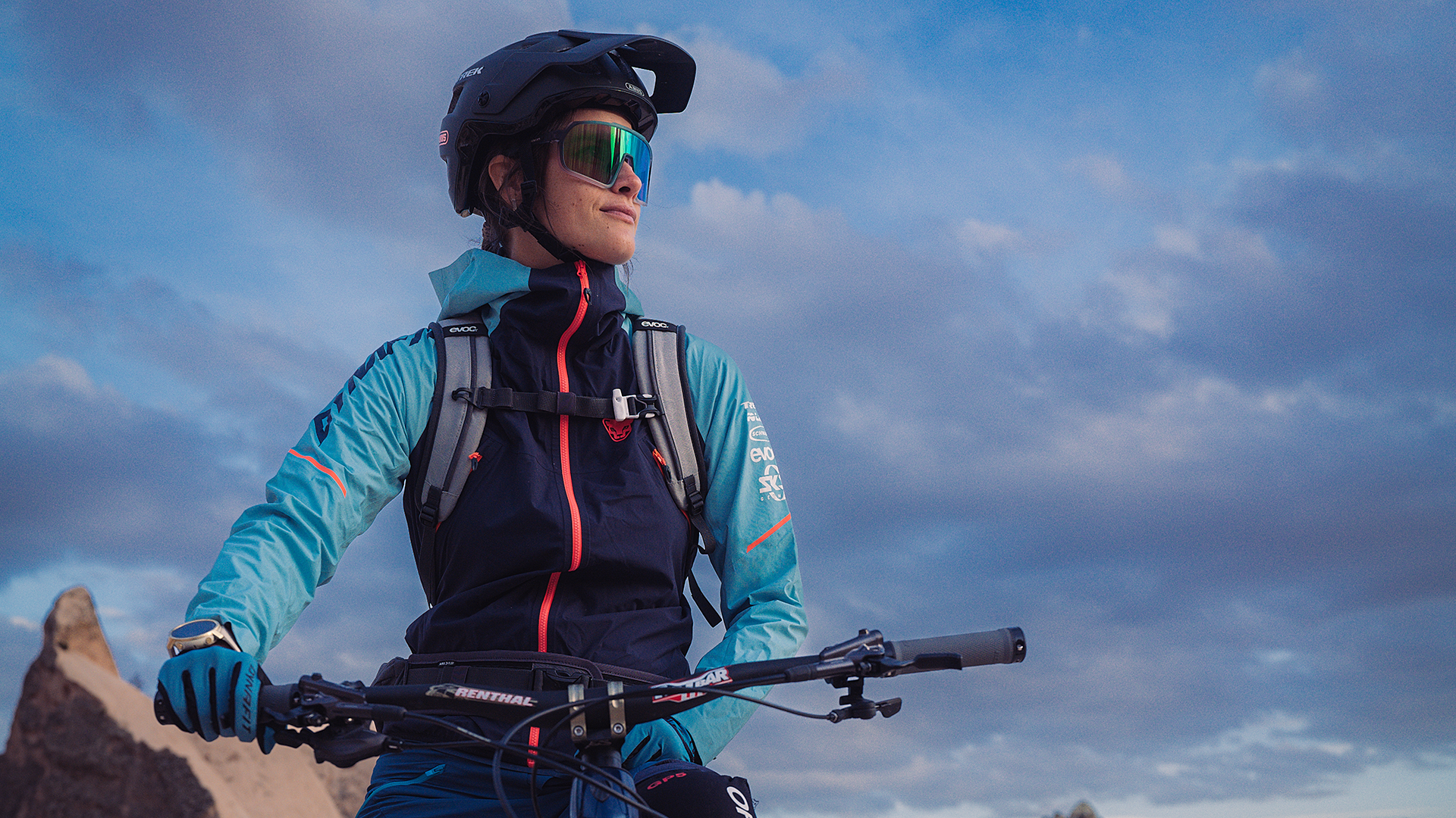 Athlete in blue and turquoise cycling jacket with cycling goggles looks at the mountain backdrop